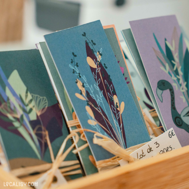 Un présentoir de cartes de vœux colorées avec des motifs de plantes et d’oiseaux, exposées dans un support en bois au magasin "Rawet" à Angleur.