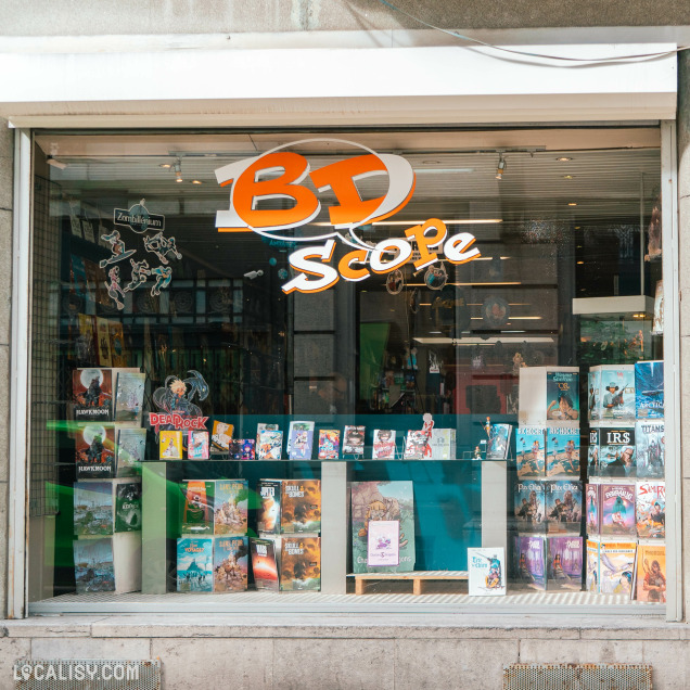 Devanture de la librairie “BDscope” à Liège. Le nom du magasin est affiché en grandes lettres orange au-dessus de la vitrine, qui expose une variété de bandes dessinées et de romans graphiques colorés.