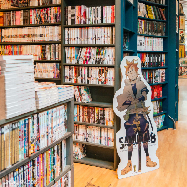 L’intérieur de la librairie “BDscope” à Liège, avec des étagères remplies de bandes dessinées colorées. Un présentoir en carton du personnage “SAMURAI 8” est bien en évidence au premier plan.