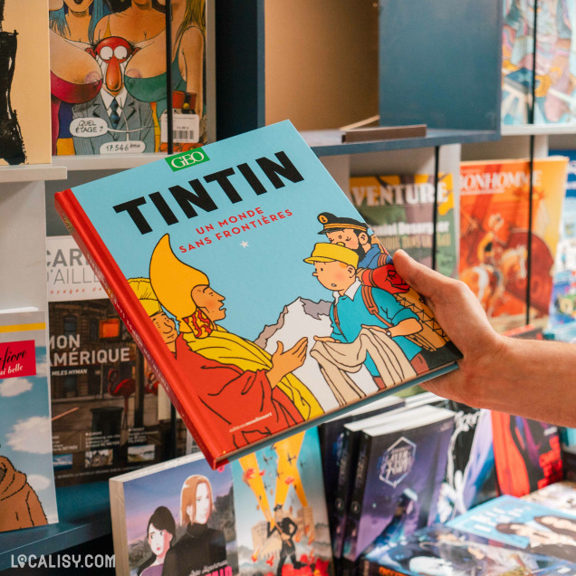 Une main tenant une bande dessinée intitulée “TINTIN UN MONDE SANS FRONTIÈRES” dans la librairie “BDscope”. En arrière-plan, on voit des étagères remplies de bandes dessinées et de romans graphiques.