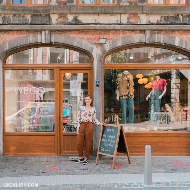 Devanture du magasin “WeCo Store” à Liège. La façade comporte une grande fenêtre en verre et une porte en verre avec des cadres en bois. Un panneau en ardoise devant le magasin indique qu’il est ouvert, et divers articles de vêtements sont exposés dans la vitrine. Une personne se tient devant la porte.