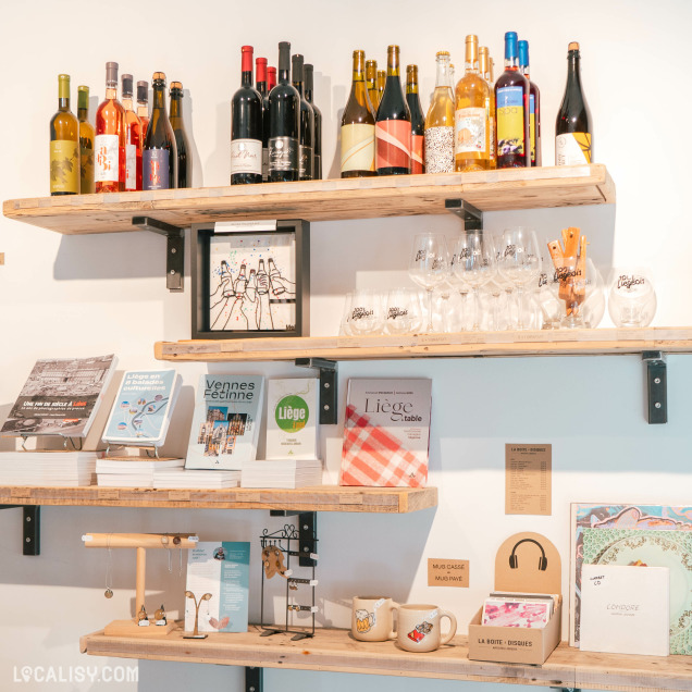 Un mur intérieur du magasin “100% Liégeois” à Liège avec trois étagères en bois exposent divers articles, principalement des bouteilles de vin et autres boissons, des verres et quelques livres.