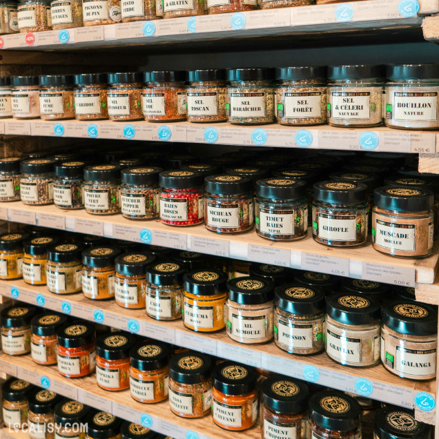 Des étagères en bois dans le magasin “100% Liégeois” à Liège, remplies de divers bocaux étiquetés avec des noms comme “Basilic,” “Sel Fin,” et “Bouillon Anisé.”