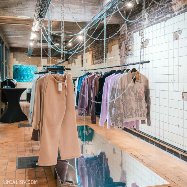 L’intérieur du magasin “Tenory” à Liège, avec des vêtements suspendus à des chaînes. Le magasin a un style industriel chic avec des murs en briques apparentes et un sol réfléchissant.