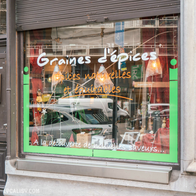 Devanture du magasin “Graines d’épices” à Liège, avec le texte “Graines d’épices” sur la vitrine. En dessous, des phrases en français indiquent que le magasin vend des épices naturelles et des tisanes, et invite à découvrir de nouvelles saveurs.