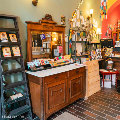 L’intérieur du magasin “Graines d’épices” à Liège, avec un comptoir central en bois exposant divers produits. Derrière le comptoir, une armoire ancienne avec des portes vitrées présente d’autres articles.