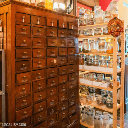 Une grande armoire en bois comportant de nombreux petits tiroirs étiquetés avec des noms d’épices. À droite de l’armoire, des étagères sont remplies de bocaux en verre contenant diverses épices à l’intérieur du magasin “Graines d’épices” à Liège.