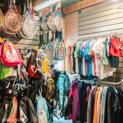 L’intérieur du magasin “Sports2life” à Embourg, avec des équipements et vêtements de sport variés. À gauche, des sacs à dos colorés et des raquettes de tennis sont suspendus. Au centre, des vêtements de sport sont accrochés sur des portants, organisés par couleur.