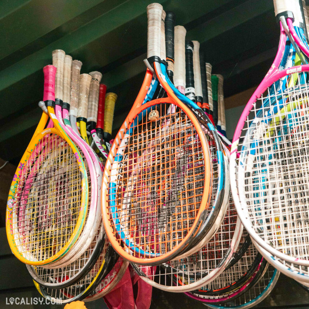 Une collection de raquettes de tennis colorées suspendues dans le magasin “Sports2life” à Embourg. Les raquettes, de différentes couleurs comme le rose, le bleu, l’orange et le blanc, sont accrochées par leurs manches, formant une disposition semi-circulaire.