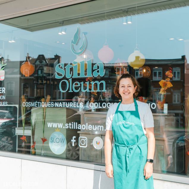 Devanture du magasin “Stilla Oleum” à Liège. La vitrine affiche le nom du magasin et un logo représentant une goutte et une feuille. Des éléments décoratifs, comme des lanternes en papier et des feuilles d’automne, sont suspendus à la fenêtre. Le texte indique que le magasin vend des cosmétiques naturels et écologiques.