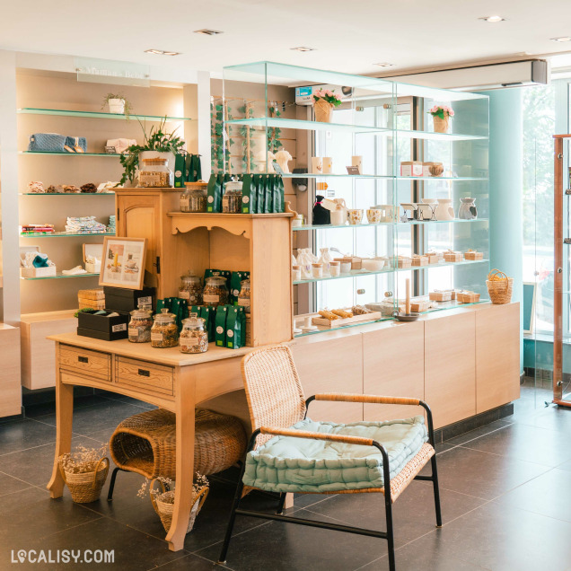 Le magasin “Stilla Oleum” à Liège présente une variété d’huiles en bouteilles et d’autres produits emballés. L’agencement est élégant et propre, avec des murs clairs, des étagères en bois et des vitrines en verre. Une table en bois avec des produits est placée au centre, accompagnée d’une chaise en osier.