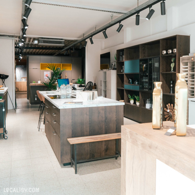 Showroom de "MyCuisine" à Neupré avec plusieurs designs exposés. Au centre, il y a un grand îlot de couleur sombre avec un plan de travail blanc, entouré de tabourets hauts. À droite, des étagères avec des portes vitrées exposent des bouteilles et des verres.