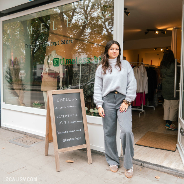 Devanture du magasin “TIMELESS Friperie” à Liège. Une personne se tient à l’entrée, partiellement cachant la vue de l’intérieur où divers vêtements sont visibles. Devant le magasin, un panneau sur le trottoir affiche le texte “TIMELESS,” “Vêtements Seconde Main,” et “@timeless_shop.”