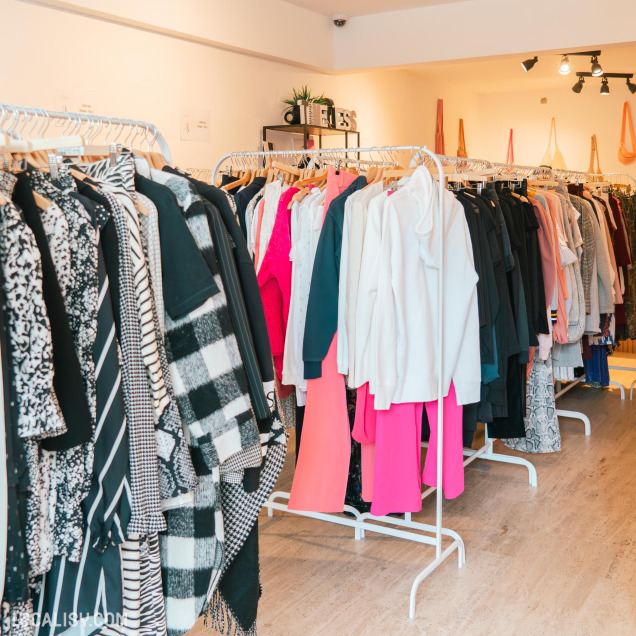 L’intérieur de “TIMELESS Friperie” à Liège, un magasin de vêtements. Plusieurs portants sont remplis de divers vêtements, y compris des manteaux, des robes et des chemises de différentes couleurs et motifs.