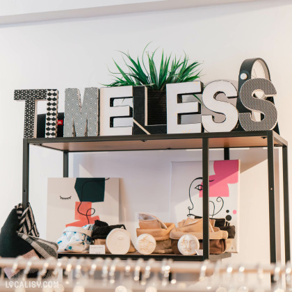 L’intérieur du magasin “TIMELESS Friperie” à Liège. Le nom “TIMELESS” est affiché en grandes lettres décoratives sur une étagère. En dessous, divers articles sont exposés, y compris des sacs, des boîtes avec des visages dessinés et ce qui semble être de petits contenants ou des bougies.