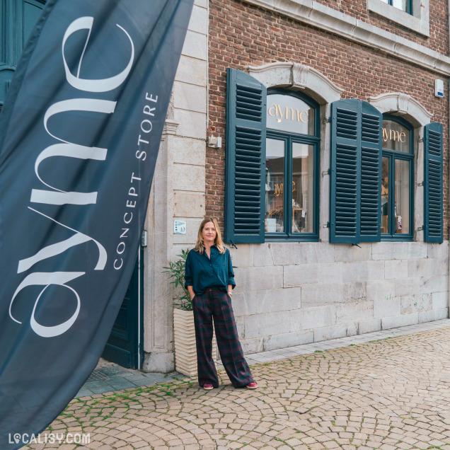 Devanture du magasin “Ayme” avec le sous-titre “Concept Store” situé à Theux. Un grand drapeau noir avec le nom du magasin en lettres blanches est placé à côté de l’entrée. Le bâtiment a une façade traditionnelle en briques avec deux fenêtres aux volets bleu foncé.