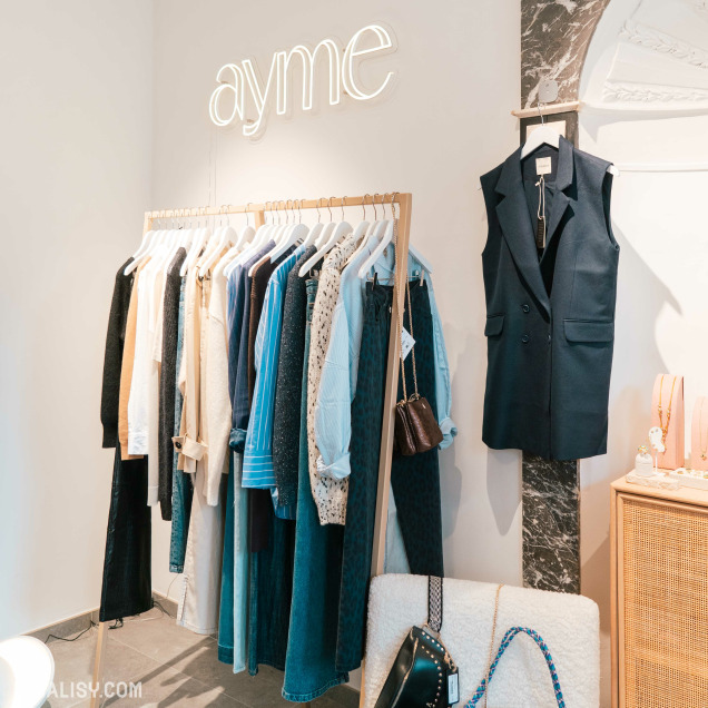 Un portant avec divers vêtements, y compris des chemises, vestes et robes de différentes couleurs et tissus chez “Ayme” à Theux. Un mannequin à droite met en valeur une veste sombre. Le décor est minimaliste avec des murs blancs à motifs de marbre et une enseigne néon “ayme” en lettres minuscules.