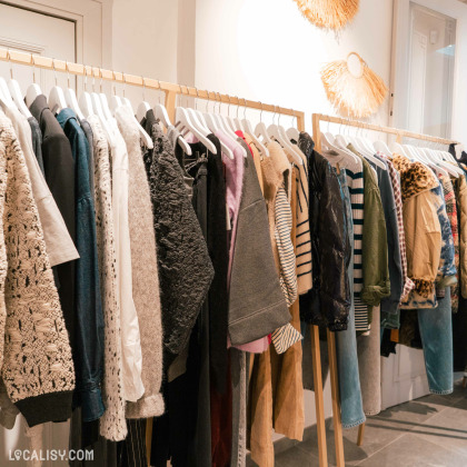 Divers vêtements suspendus sur des cintres en bois le long d’un portant en métal à l’intérieur du magasin “Ayme” à Theux. Les vêtements semblent être de la mode féminine avec différentes textures et motifs, y compris des tricots, des rayures et des couleurs unies.