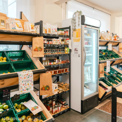 Des étagères en bois remplies de divers produits, y compris des bouteilles et des bocaux, ainsi que des produits frais exposés dans des caisses vertes. Il y a une section réfrigérée à droite avec une porte en verre, et le sol est carrelé à  l’épicerie “En fait C’est Simple” à Liège.