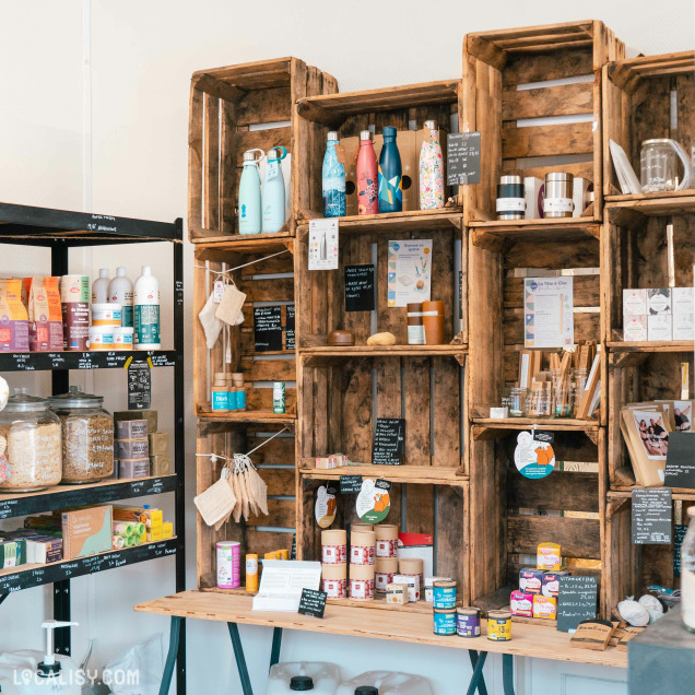 L’intérieur de l’épicerie “En fait C’est Simple” à Liège avec des étagères en bois rustiques remplies de divers produits. Il y a des bouteilles réutilisables, de la verrerie et des contenants sur les étagères supérieures, tandis que les étagères du milieu et du bas présentent des produits emballés comme des céréales et des grains.