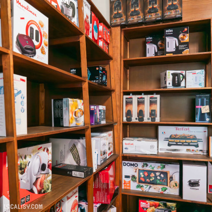 L’intérieur du magasin “Maison Ponsard” à Liège avec des étagères en bois remplies de divers petits appareils électroménagers et ustensiles de cuisine. Les marques telles que Tefal, JBL et DOMO sont visibles sur les emballages.