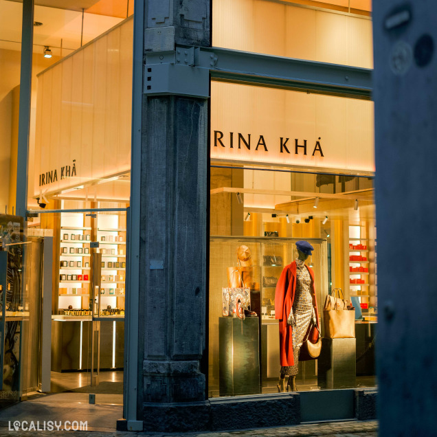 Devanture du magasin “Irina Khä” à Liège. La vitrine en verre permet de voir l’intérieur du magasin, où sont exposés divers articles sur des étagères et un mannequin vêtu d’un manteau rouge, d’un chapeau et d’une écharpe.