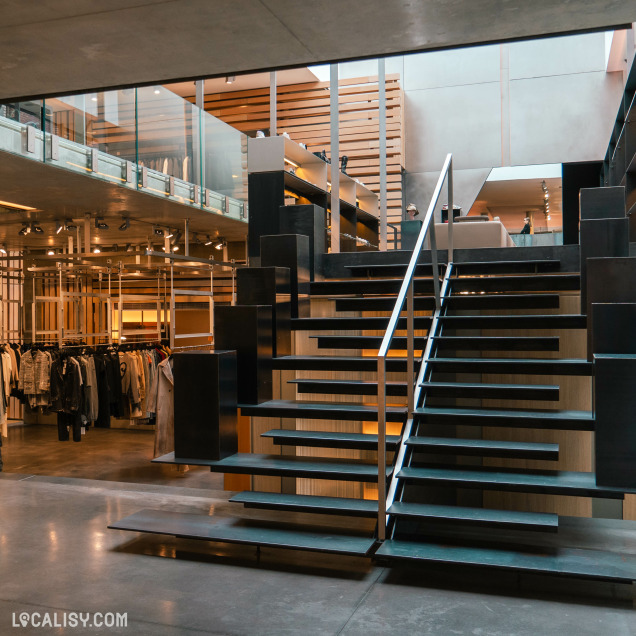 Un escalier central composé de marches noires aux bords métalliques, menant à un étage supérieur. Le magasin "Irina Khä" à Liège a une disposition ouverte avec des portants de vêtements sur les deux niveaux, exposant divers vêtements.