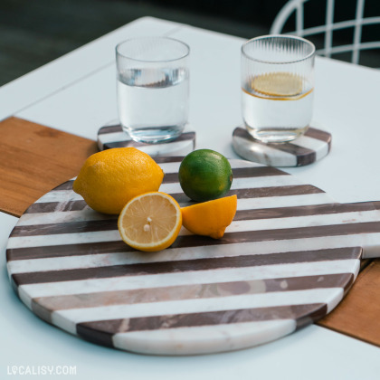 Une planche rayée en marbre est ornée de citrons et d'un citron vert, accompagnée de deux verres d'eau posés sur des sous-verres assortis, illustrant le style raffiné et moderne des articles de l'e-commerce Sista à Aywaille