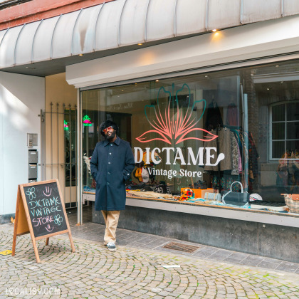 Le gérant pose devant la vitrine de Dictame, un commerce de seconde main situé à Liège, avec un panneau ardoise indiquant le nom du magasin placé à côté de l'entrée.