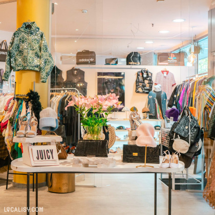 L'intérieur de Beau Bizarre à Liège présente divers vêtements et accessoires, avec une table centrale décorée de fleurs et de sacs, entourée d'articles de mode exposés sur les murs.