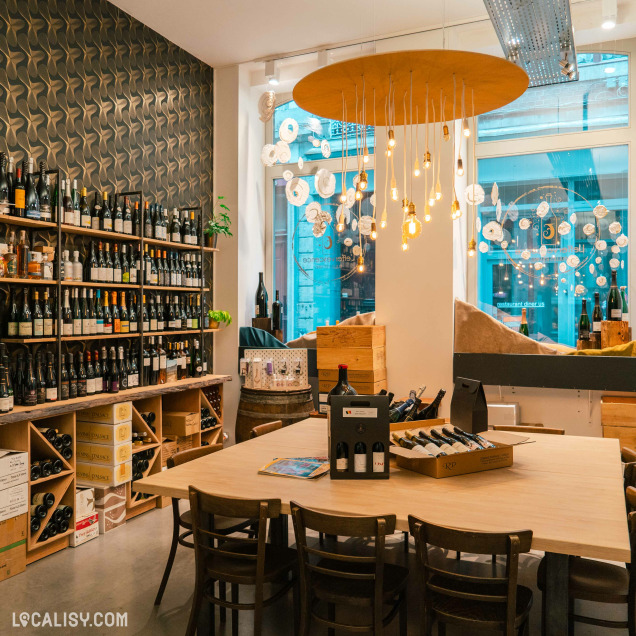 Des étagères en bois remplies de bouteilles et une table centrale pour les dégustations. La décoration chaleureuse et moderne, avec des luminaires suspendus au magasin “L’Effervescence” à Liège