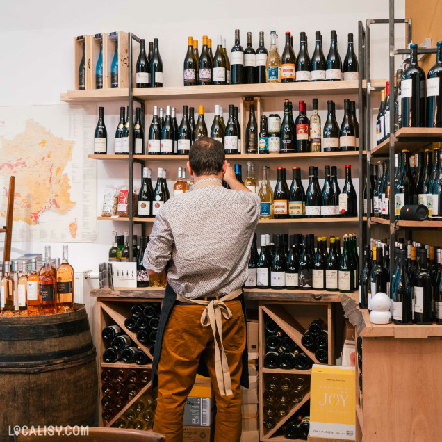 L’intérieur du magasin “L’Effervescence” à Liège avec des étagères remplies de bouteilles variées. Un tonneau en bois sert de table d’exposition, et une carte accrochée au mur ajoute une touche décorative.