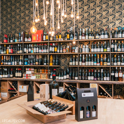 L’intérieur du magasin “L’Effervescence” à Liège présente des étagères en bois bien garnies de bouteilles de vin, avec un papier peint à motifs ondulés en arrière-plan. Des caisses en bois et un porte-bouteilles en carton sont disposés sur une table au premier plan.