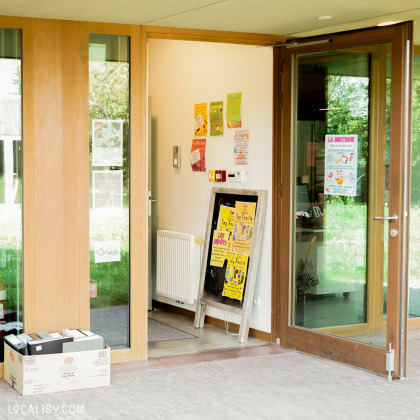 Devanture du magasin “Try’Fouille” à Herve avec des portes vitrées, dont l’une est ouverte. Des affiches et flyers sont visibles sur les portes et les fenêtres adjacentes, annonçant divers produits, services ou événements.