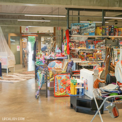 Des étagères remplies de jeux de société et de puzzles, un présentoir avec des jouets colorés pour enfants, et divers autres biens dispersés dans l’espace à l’intérieur du magasin “Try’Fouille” à Herve.