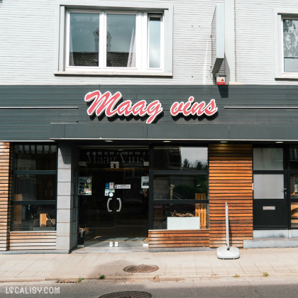 Devanture du magasin “Maag Vins,” une boutique de vins située à Visé. La façade est sombre avec de grandes fenêtres et le nom du magasin en lettres cursives rouges néon au-dessus de l’entrée.