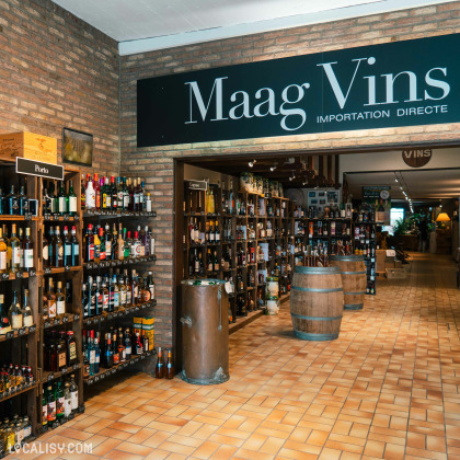 L’intérieur du magasin “Maag Vins” à Visé avec un grand panneau avec le nom “Maag Vins” en lettres blanches sur fond noir, indiquant probablement le nom de la marque du magasin. De nombreuses étagères et casiers remplis de bouteilles de vin montrent la large sélection du magasin.