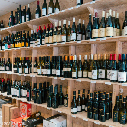 De nombreuses bouteilles de vin soigneusement rangées sur des étagères en bois qui couvrent le mur. Les étagères sont remplies de diverses bouteilles de vin au magasin “MC Vins” à Theux.