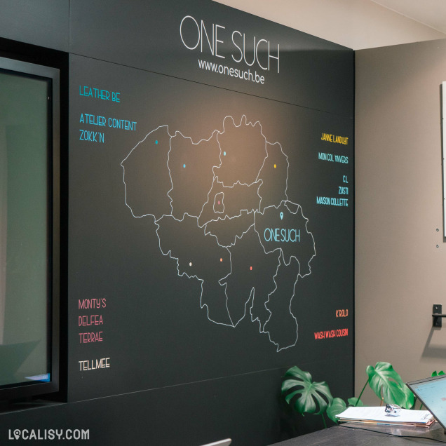 Un mur intérieur la boutique “One Such” à Liège avec un fond sombre présentant une carte stylisée de la Belgique. Sur la carte, il y a divers points colorés reliés par des lignes à des textes qui semblent être des noms de lieux ou de produits.