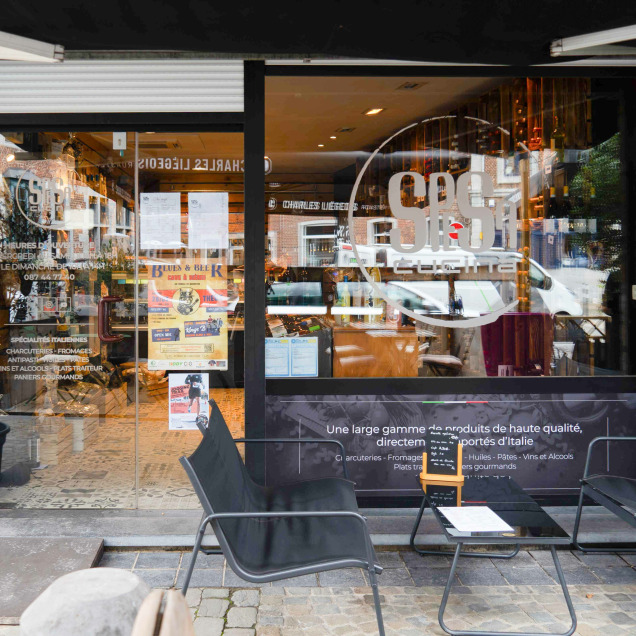 Devanture du magasin “Spesa Cucina” à Theux, la façade comporte une grande vitrine avec le nom du magasin écrit en lettres blanches. Devant la vitrine, il y a deux chaises noires et une petite table, suggérant un espace où les clients peuvent s’asseoir à l’extérieur.