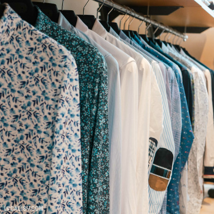 Une sélection de chemises suspendues sur un portant dans le magasin “Basile” à Ans. Les chemises présentent divers motifs et couleurs, principalement dans les tons de bleu et blanc, avec certains modèles à motifs floraux.