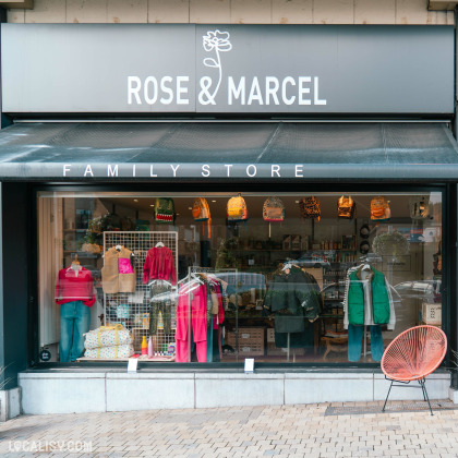 Devanture du magasin “Rose & Marcel” à Embourg. L’enseigne du magasin est noire avec des lettres blanches et comporte un graphique de rose au-dessus du texte. La vitrine expose divers articles, notamment des vêtements, des sacs et des accessoires.