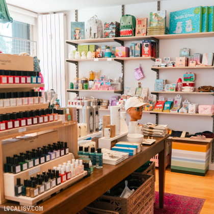 Gamme de produits exposés sur des étagères en bois et des tables au magasin “Rose & Marcel” à Embourg. Les articles incluent diverses bouteilles, probablement des cosmétiques ou des parfums, des livres, des objets décoratifs et ce qui semble être des jouets ou accessoires pour enfants.