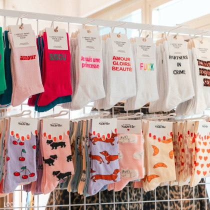 Une collection de chaussettes colorées exposées sur des cintres à l’intérieur du magasin “Rose & Marcel” à Embourg. Chaque paire de chaussettes présente des motifs et des phrases en français, telles que “amour,” “bisou,” “Je suis une princesse,” “AMOUR GLOIRE BEAUTE,” et d’autres.