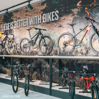 L’intérieur du magasin de vélos “53Onze Ans” à Ans. Un grand mur mural affiche le texte “LIEGE IS BETTER WITH BIKES” avec des images de cyclistes traversant une zone boisée. Devant le mur, plusieurs vélos de montagne sont exposés, dont un surélevé sur une plateforme.