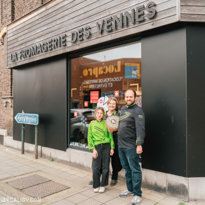 Devanture de "La Fromagerie des Vennes" à Liège. Une façade avec un auvent sombre portant le nom du magasin en lettres blanches. Une grande vitrine en verre expose divers articles et affiches. Trois personnes se tiennent sur le trottoir devant le magasin.