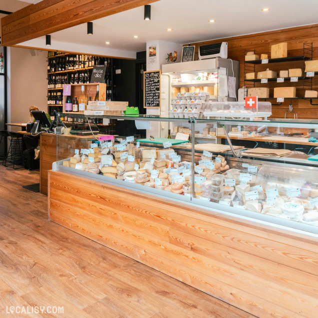 L’intérieur de “La Fromagerie des Vennes” à Liège montre un grand comptoir vitré rempli de fromages, des étagères en bois avec des produits, et un menu sur tableau noir.