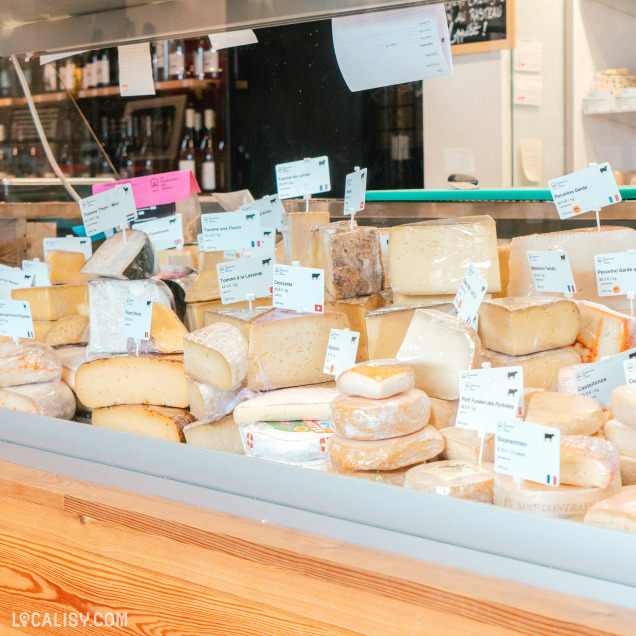 Un étalage de différents types de fromages à “La Fromagerie des Vennes” à Liège. Les fromages sont disposés sur des étagères derrière un comptoir en verre, chaque pièce ayant une étiquette d’information.