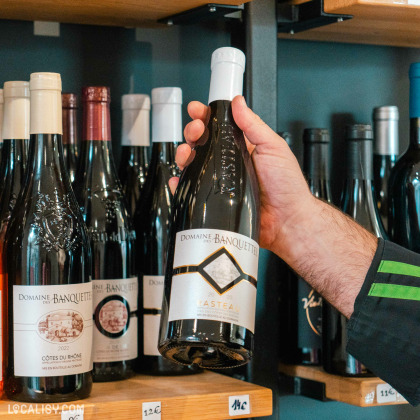 Une main tenant une bouteille de vin devant des étagères remplies de diverses autres bouteilles à “La Fromagerie des Vennes” à Liège. L’étiquette indique “Domaine Banquet”.