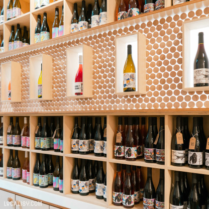 Une étagère murale remplie de bouteilles de vin. Les étagères en bois clair ont des découpes circulaires pour maintenir les bouteilles, formant un motif en nid d’abeille à l'intérieur du magasin “Les Vintrépides” à Liège.